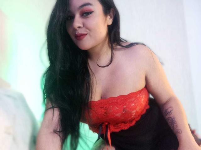 Ambeerx69 con lunghi capelli neri, una camicetta di pizzo rossa che mostra il seno, un pantaloncino nero, un tatuaggio sul braccio destro, uno sfondo verde e bianco e un sorriso sicuro..