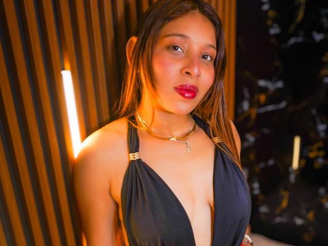 JaneKetty with wet brown hair, red lipstick, black deep-cut top showing her small tits, gold chain necklace, and warm lighting. She's looking at the camera with a slight smile.