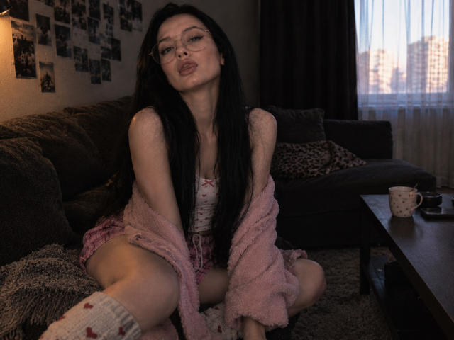 Alanna Orsburn, avec de longs cheveux noirs, des lèvres pulpeuses, des lunettes, une veste rose duveteuse portée en bandoulière, un débardeur blanc, un short rose, des chaussettes, accroupie sur un canapé sombre, dans une pièce faiblement éclairée, avec vue sur la ville..