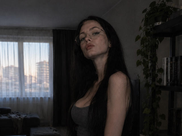 Alanna Orsburn, met lang zwart haar, een bril en een grijze tanktop, waarbij haar borsten zichtbaar zijn, kijkt naar beneden met een lichtjes fronsende gezichtsuitdrukking in een slecht verlichte kamer..