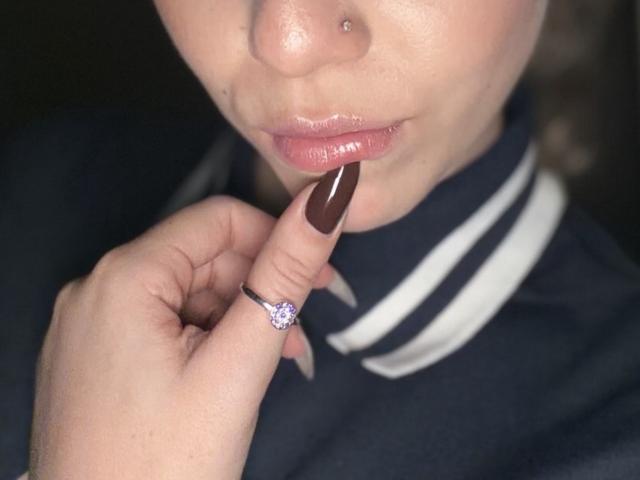 Le nez de SugarrX présente un petit piercing, légèrement obstrué. Elle mordille son ongle brun, manucuré, et montre une bague avec un joyau violet. Ses lèvres roses et brillantes sont légèrement entrouvertes..