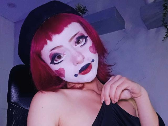 ViolettClay with red makeup, white face, heart and black lip patterns, wearing a black hat, pulling down a black strap, showing bare shoulders, red hair, and purple eyes.