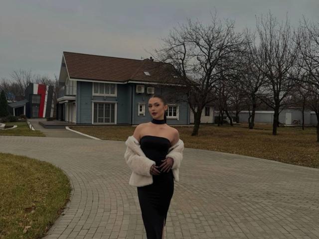 SofiiKiss se tient devant une maison, vêtue d'une robe noire sans bretelles, d'une veste en fourrure blanche et de gants violets, avec des arbres nus et un ciel gris et nuageux..