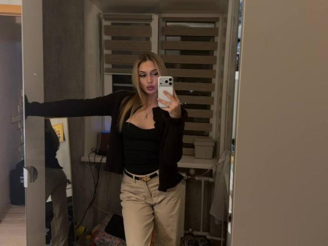 SofiiKiss, avec de longs cheveux blonds, un haut noir, une veste marron et un pantalon beige, prend un selfie devant un miroir, montrant ses seins, tout en tenant un téléphone blanc, dans une pièce peu éclairée..