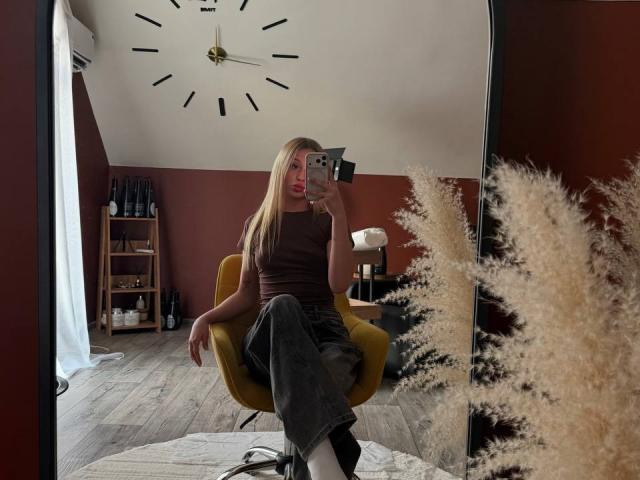 SofiiKiss com cabelos longos e loiros, blusa marrom sem mangas, calças cinzas, cadeira amarela, segurando o celular, selfie no espelho, penugem branca e fofa em primeiro plano, piso de madeira, parede vermelha..