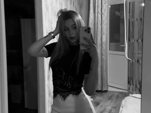 Black-and-white mirror selfie of SofiiKiss with long hair, wearing a lace top and white pants, holding a phone, hand in hair, visible ass, wooden floor, and curtains.