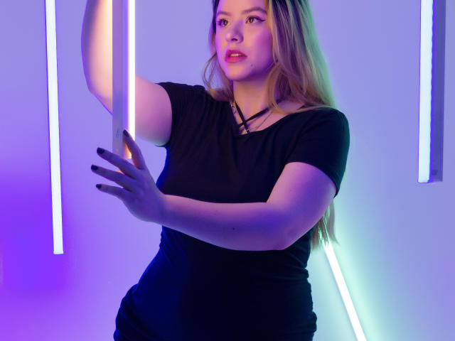 AshleySofia with long blonde hair, wearing a black top, touching a neon purple and blue strip with her right hand, looking slightly to the side with a soft expression.