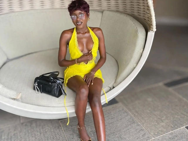 EbonySweet-hot with dark brown skin and short red hair, wearing a yellow lace halter dress, sitting on a white wicker chair, legs crossed, black handbag on the seat.