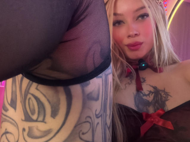 MiaValenttine with blonde hair, red lipstick, and a black choker, looking at a tattooed leg in sheer black stockings, showing her tits in a black top with a red bow.