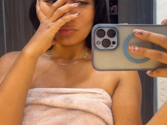 AmeliRoux with tan skin, black hair, covering her nose with her hand, holding a phone, wearing a pink towel over her tits, and a delicate necklace.