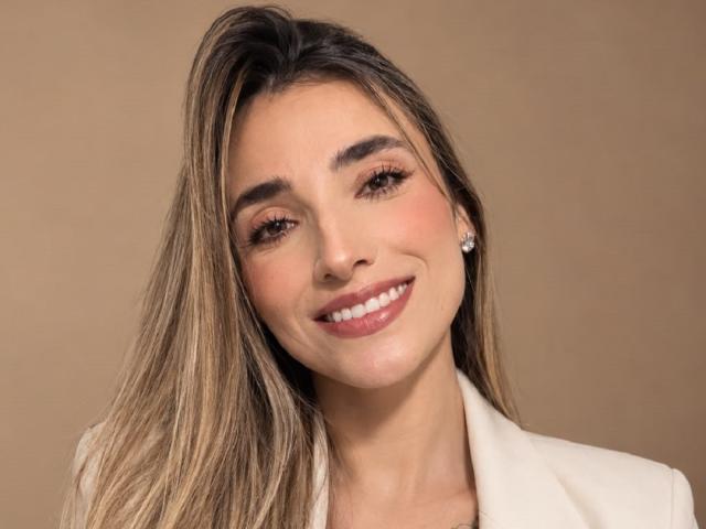 AdharaCooperr ha lunghi capelli castani, un trucco leggero, rossetto rosa e un blazer bianco. Sorride, indossa piccoli orecchini di diamanti e i suoi seni sono leggermente visibili sotto il blazer..