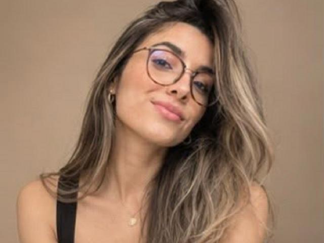 AdharaCooperr with long, wavy brown hair, wearing glasses, black strap, and a subtle smile, has pink lipstick and a small gold necklace, against a beige background.
