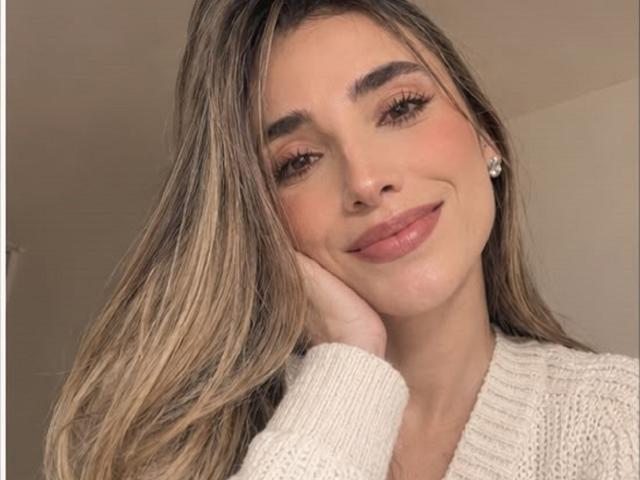 AdharaCooperr with long blonde hair, brown eyes, and pink lips, wearing a white knitted sweater, smiling softly with hand on cheek, wearing small diamond earrings.