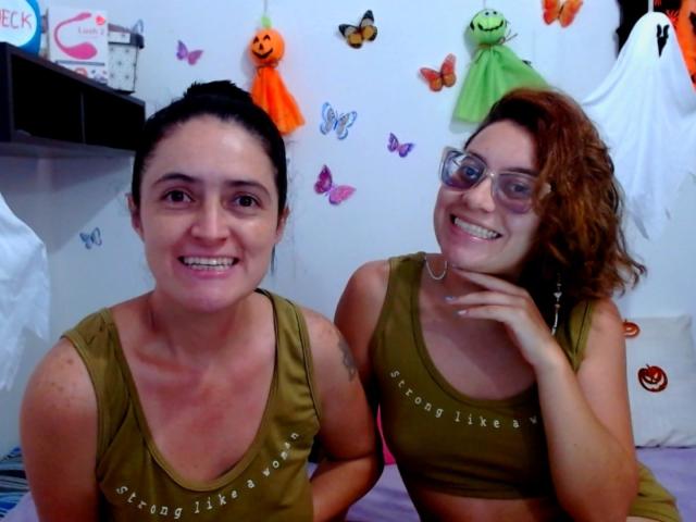LunaVenus with short dark hair and LunaVenus with curly brown hair, both wearing olive green sports bras, smiling at the camera, showing their tits, in a brightly decorated room.