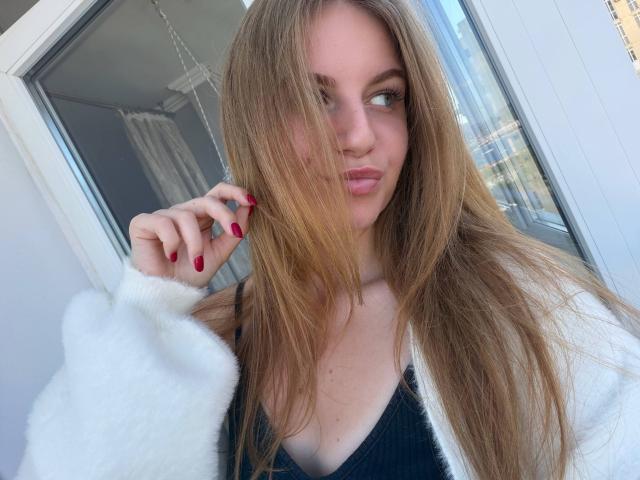 Nanette Dawley, con i suoi lunghi capelli biondo scuro, le unghie rosse, le labbra piene e un cardigan bianco soffice sopra una maglietta nera, guarda di lato con un'espressione leggermente infastidita..