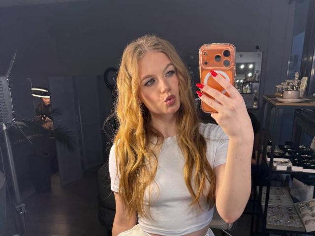 NanetteDawley with long blonde wavy hair, white t-shirt, red nails, pouting lips, holding orange phone, taking selfie, showing her tits, in dimly lit room.