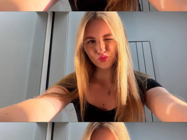 Nanette Dawley, avec de longs cheveux blonds, clignant de l'œil, lèvres boudeuses, portant un haut noir, un collier, prenant un selfie, bras légèrement tendus, lumière venant du côté, teint de peau chaud..