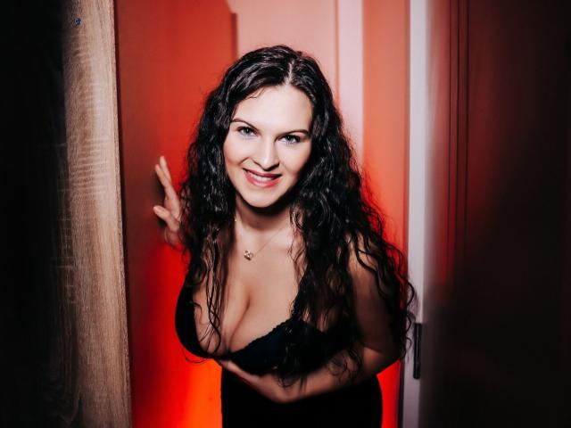 Kathy Cole, con i capelli scuri e ondulati, sorridente, indossa un reggiseno di pizzo nero, mostra il seno, tiene aperta una porta, con una luce rossa sullo sfondo, fa il gesto della mano "rock on", indossa una collana, è in piedi nell'ingresso..