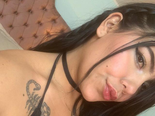 CatalinaReiyna with black hair, brown eyes, and pink lips, showing off a snake tattoo on her left boob, wearing a black choker. Soft smile, slight nose piercing.