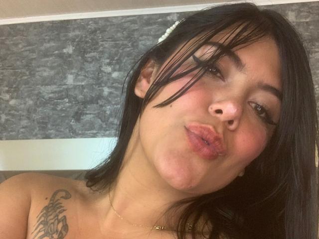 Catalina Reiyna, con capelli neri, labbra carnose, un piccolo piercing al naso e un tatuaggio sulla spalla destra, indossa una sottile collana e guarda verso la fotocamera. Il seno è parzialmente visibile, sullo sfondo si vede una parete grigia con una texture..