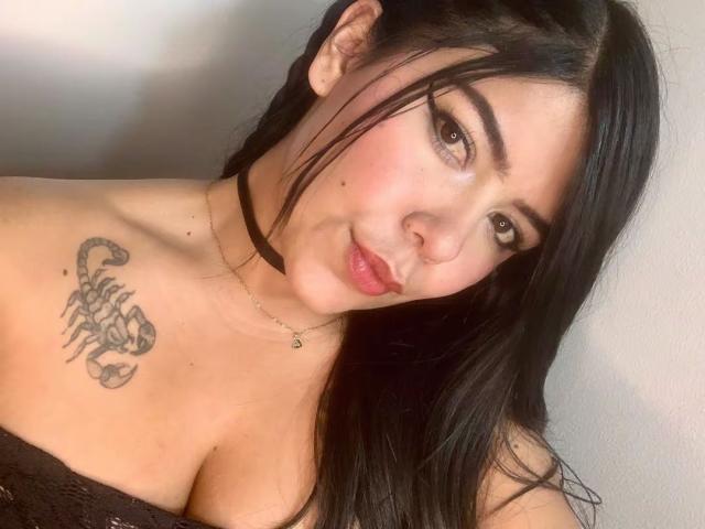 CatalinaReiyna with black hair, brown eyes, and a scorpion tattoo on her shoulder, wearing a black choker and a lace top, showing her tits and a slight smile.