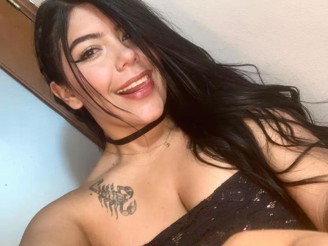 Catalina Reiyna, con i suoi lunghi capelli neri, sorride, mostra la lingua, indossa una camicetta nera in pizzo che rivela il seno e una collana nera. Ha un tatuaggio sulla spalla destra..