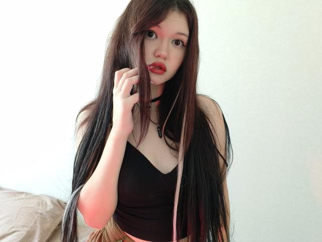 MistyEllstrom has long black hair, red lipstick, and a black top, holding her hair with a hand, showing her pale skin and small breasts, against a white wall.