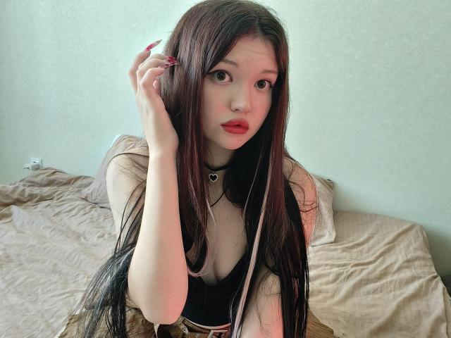 MistyEllstrom with long brown hair, wearing a black top, heart choker, and red lipstick, touching her hair with red nails, sitting on a bed with beige sheets.