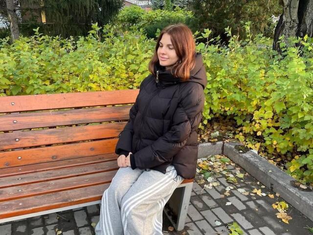 CicelyDence, with red hair, sits on a wooden bench, wearing a black puffy jacket and grey track pants, looking down with a slight smile. Her ass is partially visible.