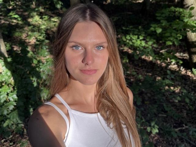 ReginaRosse has blue eyes, long straight blonde hair, and is wearing a white tank top with spaghetti straps, showing her small tits and a subtle smile.