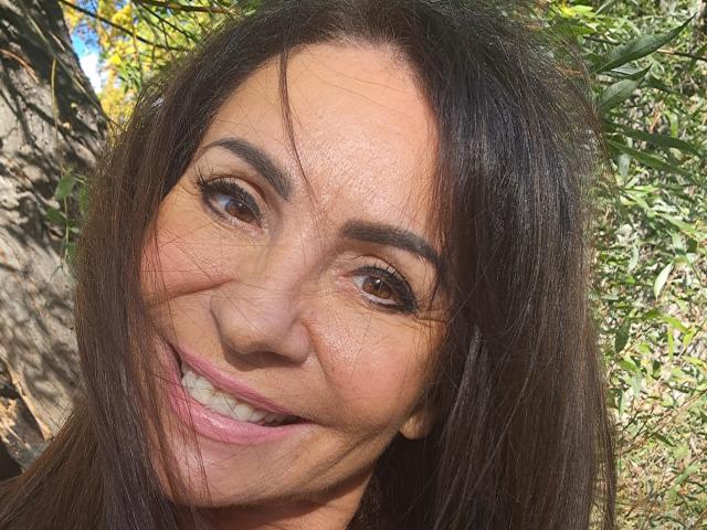 AlizeeSensual's face, showing brown eyes, dark brown hair, slight smile, and visible teeth, with a few strands of hair in her face, outdoors in sunlight.