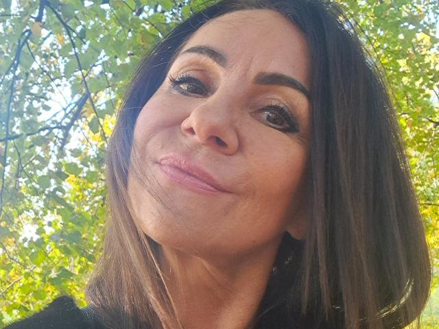 A AlizeeSensual tem uma pele lisa e marrom, cabelos longos e castanhos, e está sorrindo com lábios rosados levemente franzidos. Ela tem sobrancelhas escuras, cílios longos e está olhando para cima..