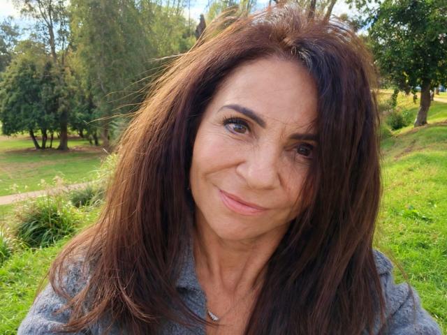 AlizeeSensual heeft bruin haar, een lichte glimlach en draagt een grijze blouse met een ketting. Ze kijkt met bruine ogen in de camera, in een zonnig, groen park..