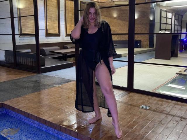 AvaQueen, a blonde woman with medium tits, wears a black sheer robe and exposes her right thigh, standing by a blue-tiled pool, smiling, with wet wooden floor.