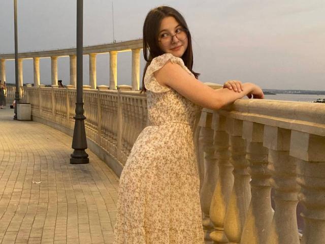 AgripinaKapke with medium tits, wearing a floral dress, leaning on a stone railing, smiling, brown hair, glasses, on a brick-paved waterfront, dusk, holding railing with hands.