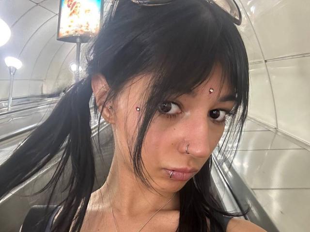 TrenaShad with black pigtails, multiple facial piercings (nose, lip, and above her lip), and black eyeliner, in an underground subway, looking at the camera.