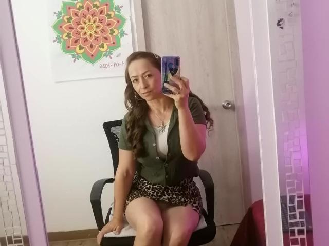 GianineLarose, con cabello largo y castaño, camisa verde de botones y falda de estampado de leopardo, sentada en una silla negra, sosteniendo un teléfono, selfie tomada con un espejo, puerta de madera, pared blanca y un colorido póster de mandala..