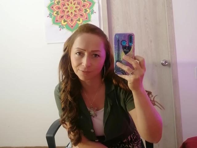 GianineLarose, con cabello castaño largo, una camisa verde, una blusa blanca, aros grandes y un collar, tomando una selfie en el espejo con una funda de teléfono morada, sonriendo ligeramente, mostrando su trasero..