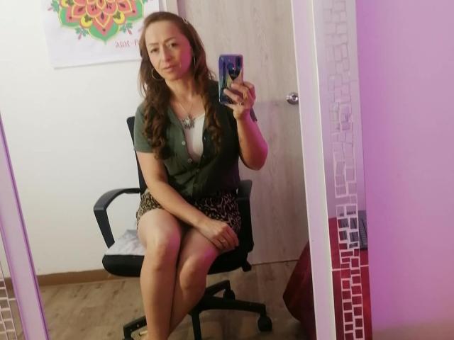 GianineLarose sentada en una silla de oficina negra, vistiendo una blusa verde y una falda de estampado de leopardo, tomándose un selfie frente al espejo con su teléfono, mostrando sus pechos y glúteos..