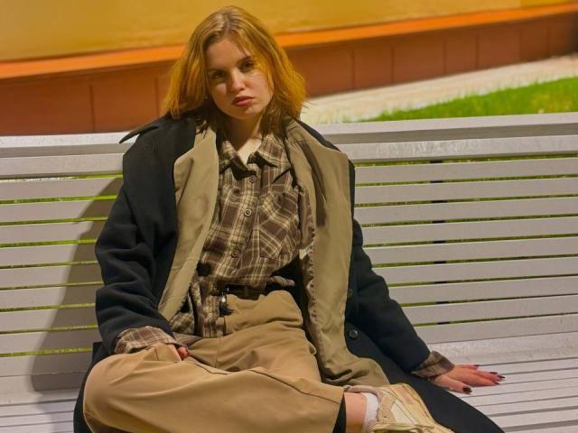 TabathaMackenthun with red hair, wearing a plaid shirt, beige pants, and a black coat, sitting on a gray bench, looking pouty, with tan sneakers, in a nighttime shot.