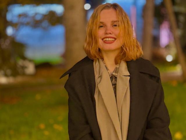 TabathaMackenthun with shoulder-length blonde hair, smiling, wearing a black coat and beige scarf, standing outdoors at night, showing her ass in the background blur.