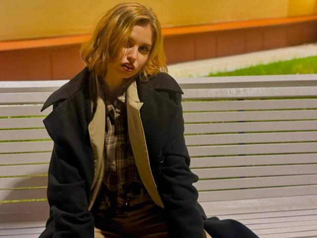 TabathaMackenthun with messy blonde hair, pouty lips, wearing a black coat over a plaid shirt, sitting on a white bench at night, looking slightly annoyed.