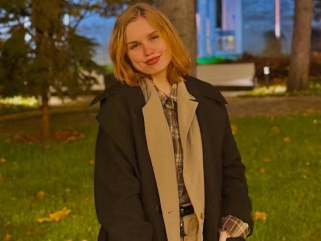 TabathaMackenthun has blonde hair, a cute smile, and is wearing a black coat over a beige and plaid shirt, standing on a grassy area with trees in the background.