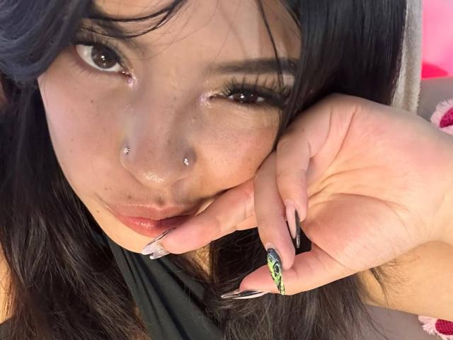 LiliMilk with black hair, brown eyes, and shiny makeup, biting her finger with long, green and black manicured nails, nose piercings, and glossy lips, looking seductively at the camera.