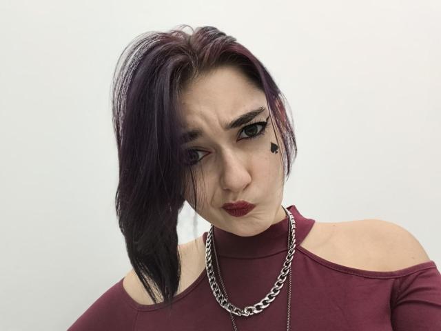 Ligia Gottshall heeft donkerpaars haar, zwarte eyeliner, rode lippenstift, een kleine zwarte harttatoeage onder haar rechteroog, en draagt een donkerrode top met een open schouder en een zilveren ketting..