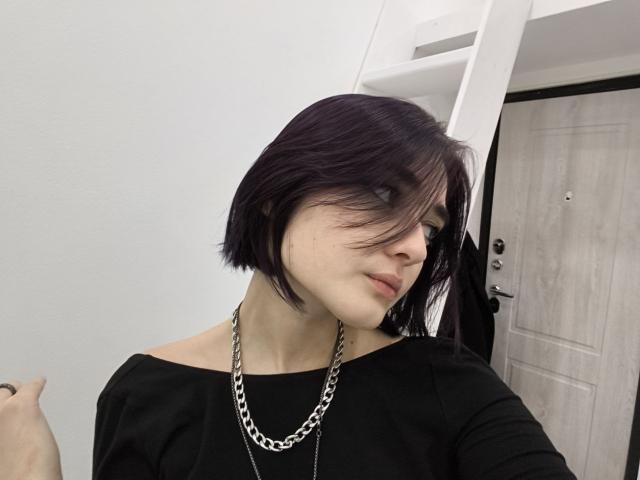 Ligia Gottshall heeft een kort, paars kapsel, bleke huid en draagt een zwarte top met een open schouderlijn, met een zilveren ketting, en kijkt schuin met een lichte glimlach..