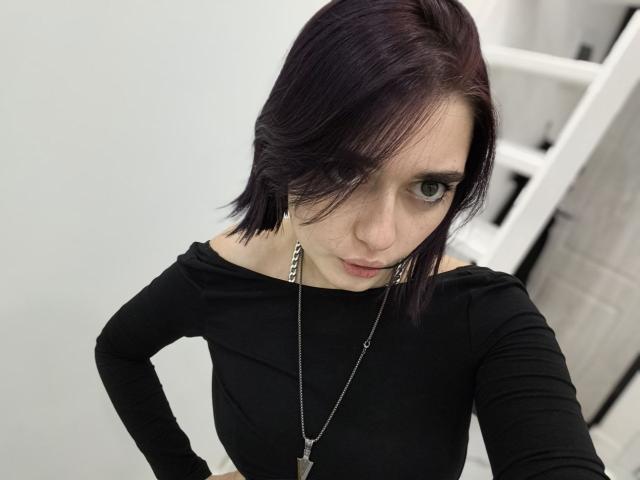 LigiaGottshall with short purple hair, black off-shoulder top, silver necklace, and serious expression, standing with hands on hips, green eyes looking up.