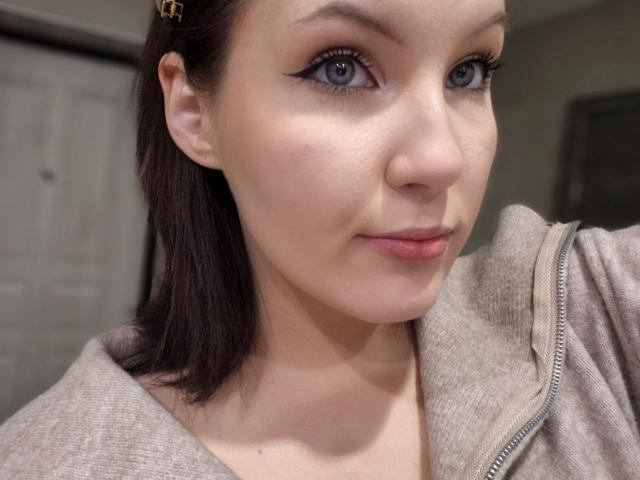 LidiaOckimey has fair skin, blue eyes with black eyeliner, brown hair in a clip, and is wearing a beige zip-up jacket, smiling subtly. Her tits are not visible.