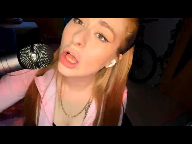 MarylnMancinelli with long brown hair, pink jacket, and gold necklace, holding a microphone, mouth open, wearing white earbuds, looking up with eyeliner and pink lipstick.