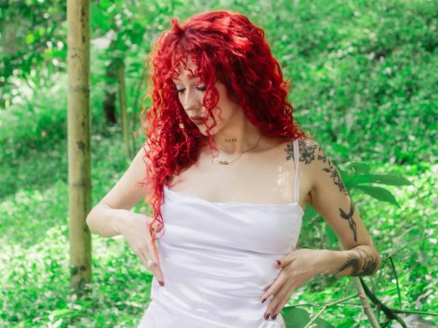 GaiaDoll com cabelo ruivo encaracolado, vestido branco sem alças, braços tatuados e mãos sobre os seios, em pé em uma floresta verde, olhando para baixo..